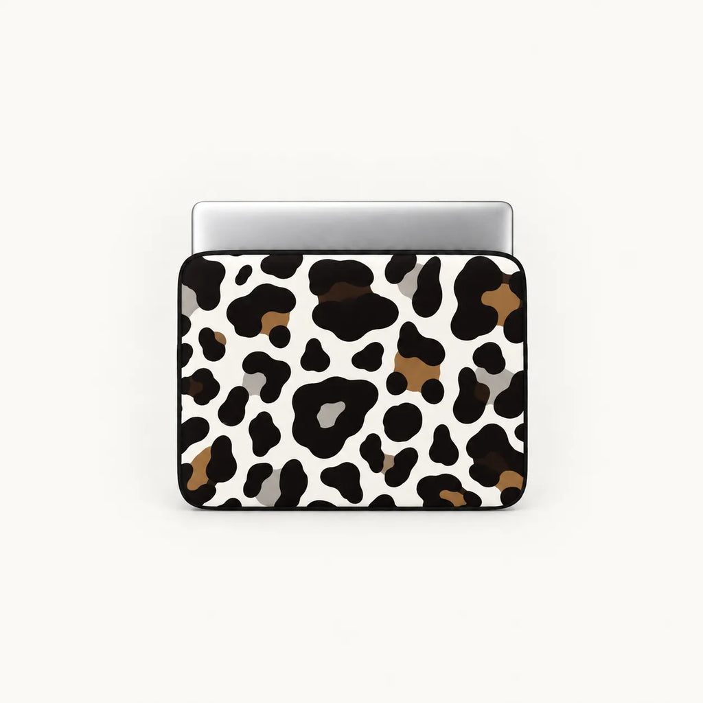 A laptop sleeve featuring a cow print pattern in black, brown, and gray on a white background, shown with a silver laptop on top against a light off-white background.