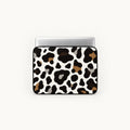 A laptop sleeve featuring a cow print pattern in black, brown, and gray on a white background, shown with a silver laptop on top against a light off-white background.