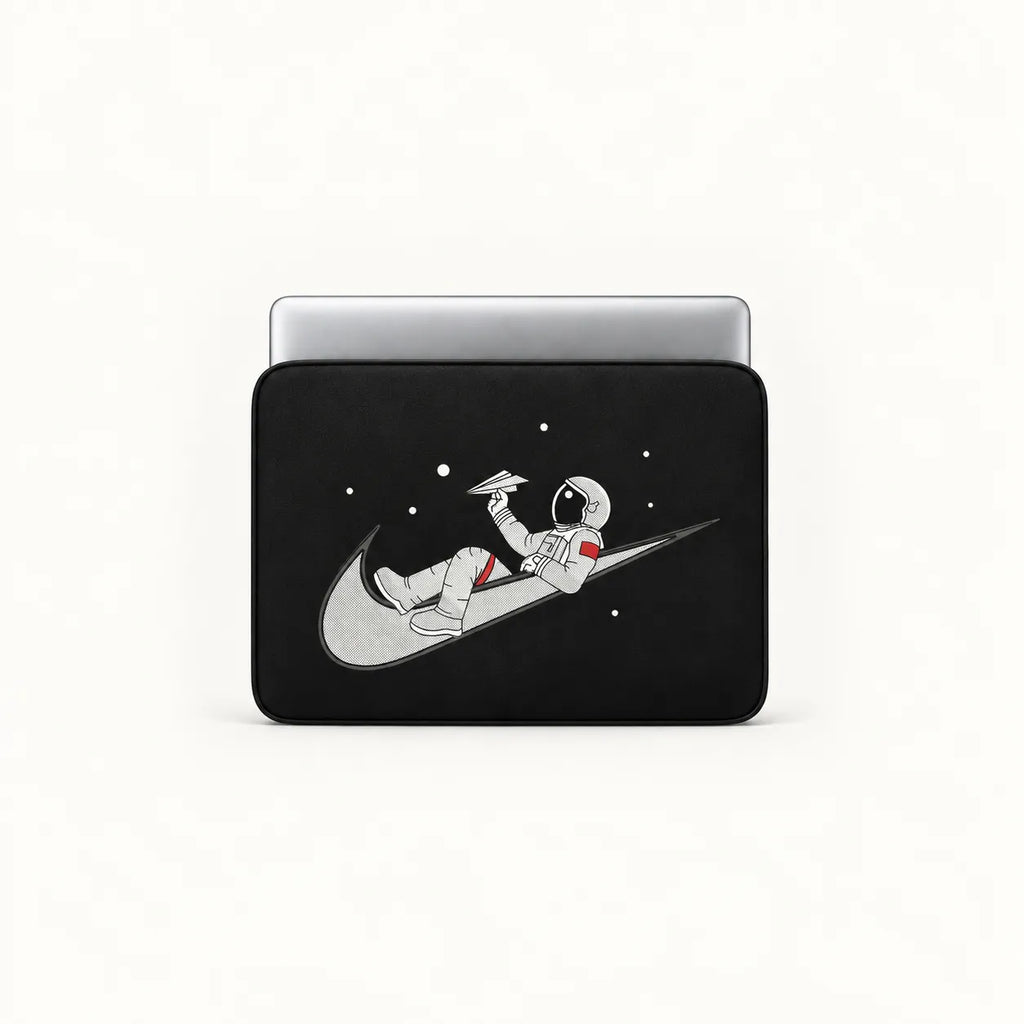 A black laptop sleeve featuring a graphic of an astronaut floating on a gray Nike swoosh against a black space background with white stars, shown on a silver laptop against a white studio background.