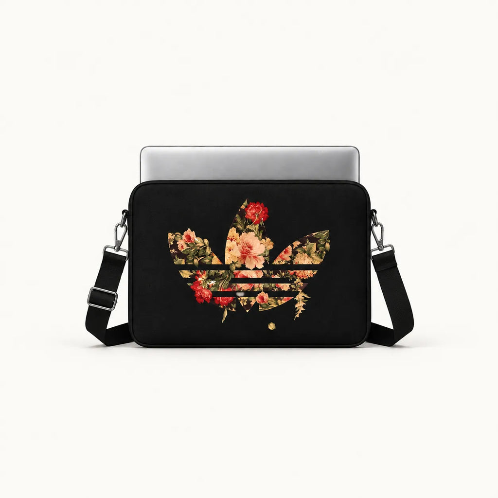 A black crossbody laptop bag featuring a floral trefoil logo design on the front, shown with a silver laptop inserted. The bag has an adjustable black shoulder strap and is photographed against a clean, off-white background.