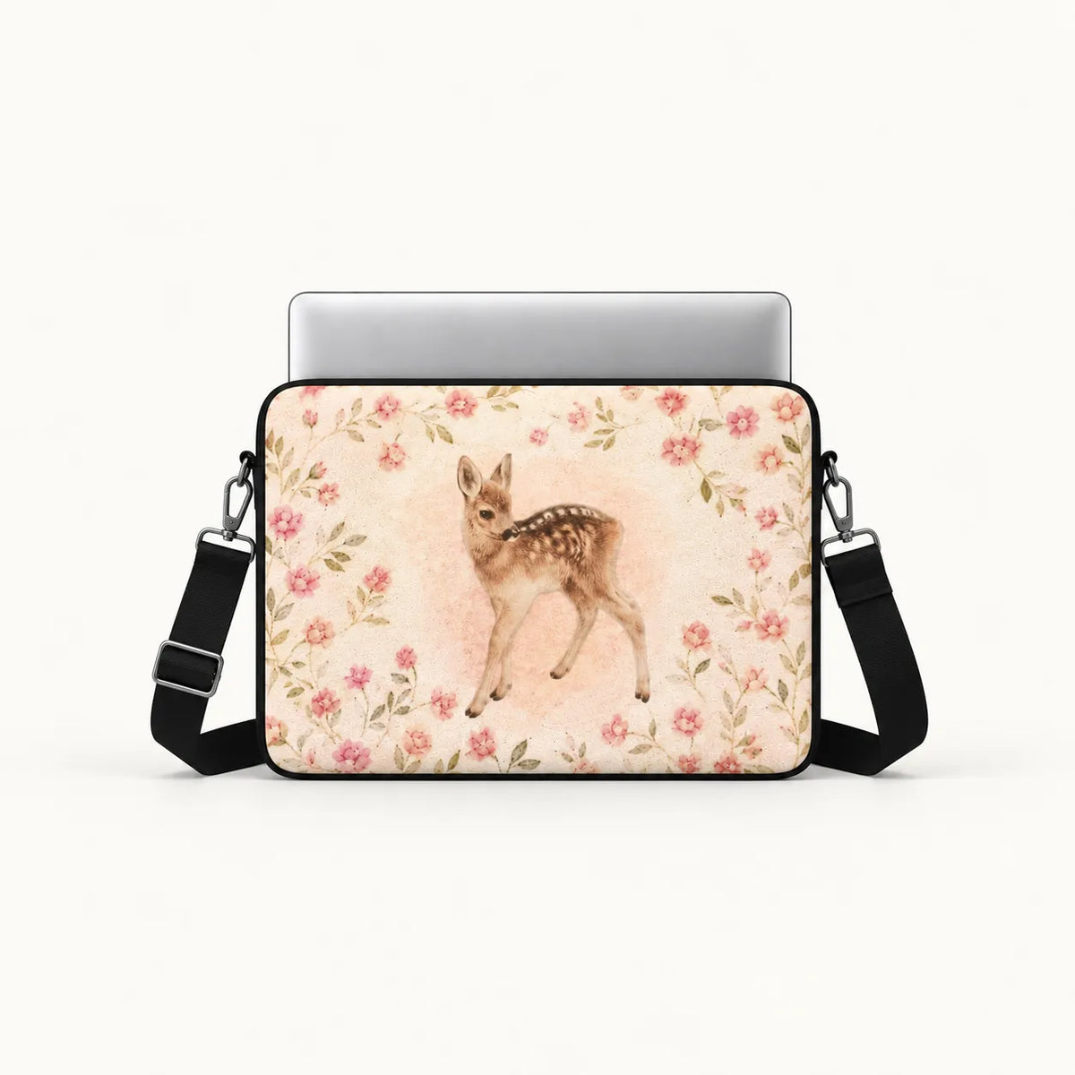 A laptop sleeve featuring a watercolor-style fawn surrounded by pink and red flowers on a cream background, shown with a silver laptop and black shoulder strap against a light gray studio background.