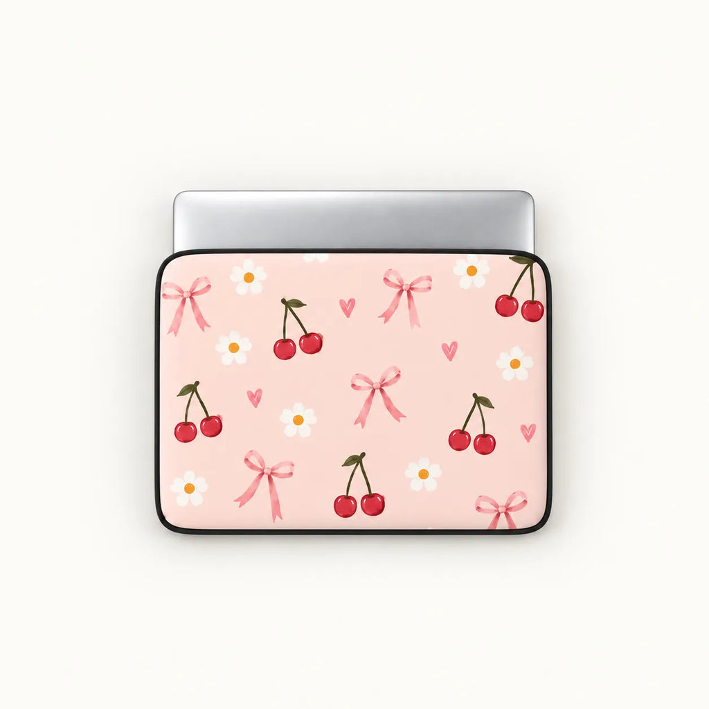 A laptop sleeve with a light pink background, decorated with a pattern of red cherries, white daisies, pink bows, and small pink hearts, shown on a silver laptop against a light off-white background.
