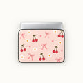 A laptop sleeve with a light pink background, decorated with a pattern of red cherries, white daisies, pink bows, and small pink hearts, shown on a silver laptop against a light off-white background.