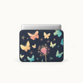A laptop sleeve with a dark navy blue background, decorated with a pattern of pastel-colored butterflies, dandelions, and small floral elements, shown with a silver laptop inserted. The product is photographed against a clean, off-white background.
