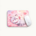 A rectangular mouse pad with a pastel illustration of a sleeping gray cat wearing a pink bow, surrounded by pink hearts and a crescent moon on a pink background. A white computer mouse rests on the right side of the pad against a light off-white background.