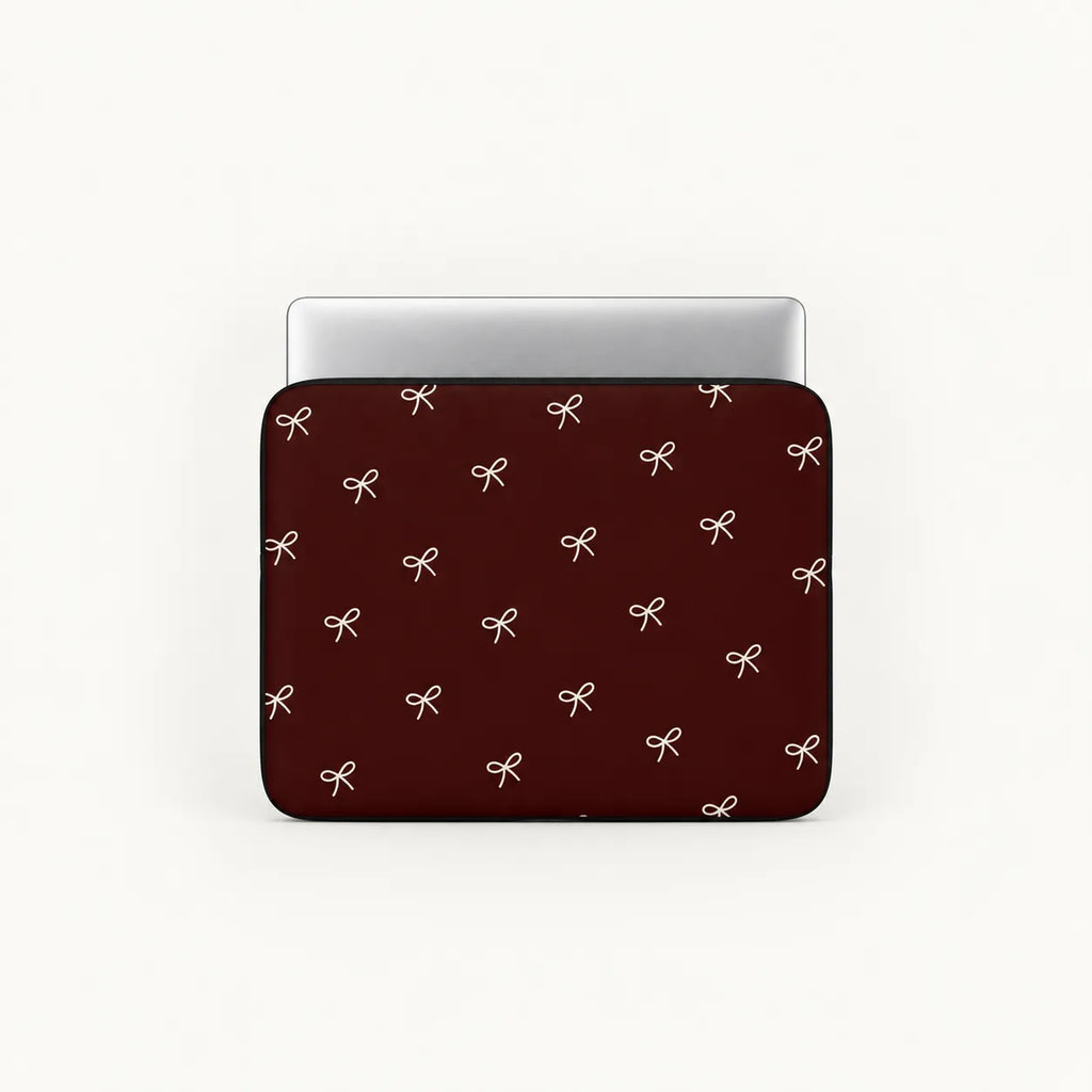 A laptop sleeve with a dark red background and a repeating pattern of small white bows, shown with a silver laptop inserted on a light gray background.