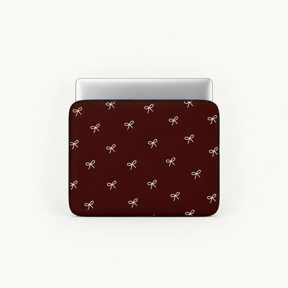 A laptop sleeve with a dark red background and a repeating pattern of small white bows, shown with a silver laptop inserted on a light gray background.
