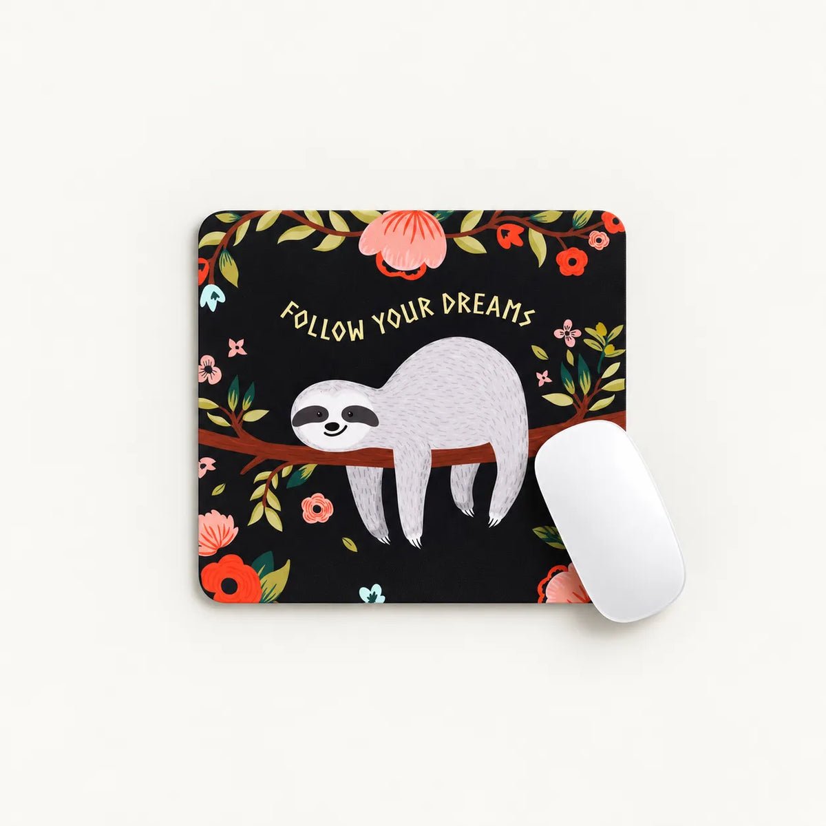 A square mouse pad with a black background featuring a gray sloth on a branch, surrounded by colorful flowers and leaves, with the text 'FOLLOW YOUR DREAMS'. A white computer mouse rests on the right side of the pad against a light off-white background.