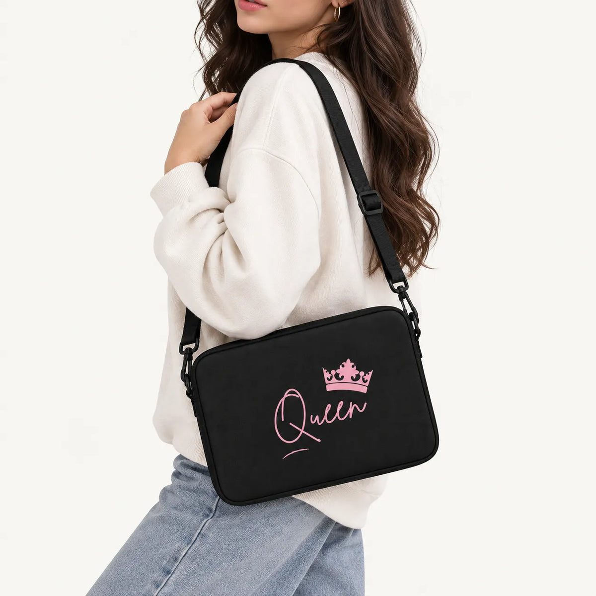 A woman wearing a white sweatshirt and light blue jeans carries a black crossbody bag with pink 'Queen' script and a crown graphic on a plain white background.