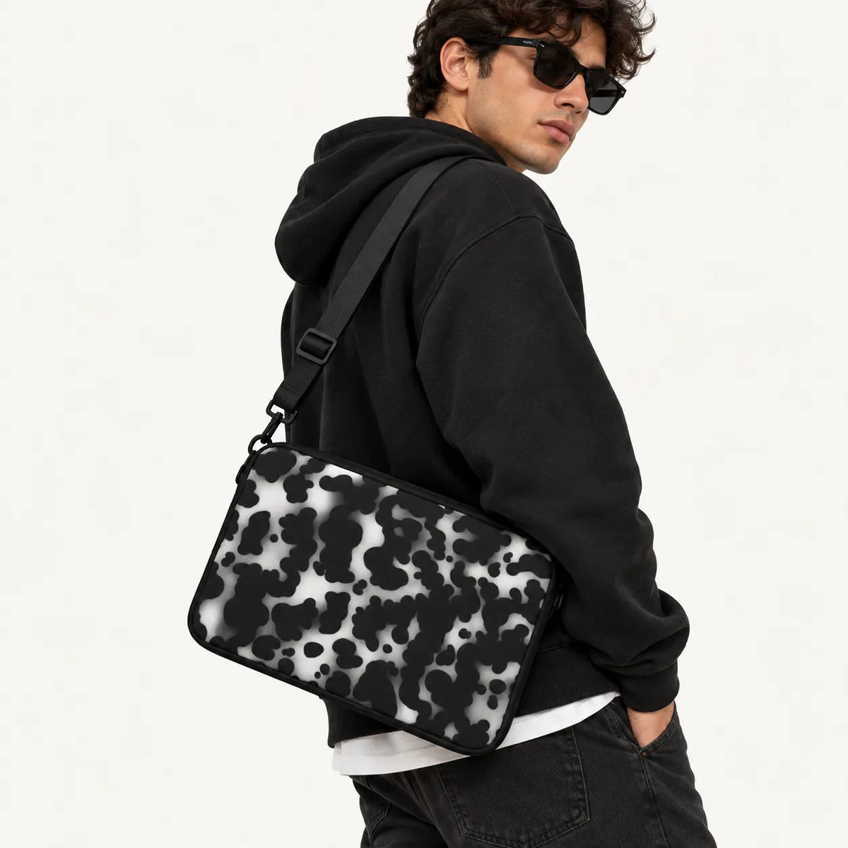 A man wearing a black hoodie and sunglasses carries a black and white cow-print shoulder bag over his shoulder, photographed against a clean white background.
