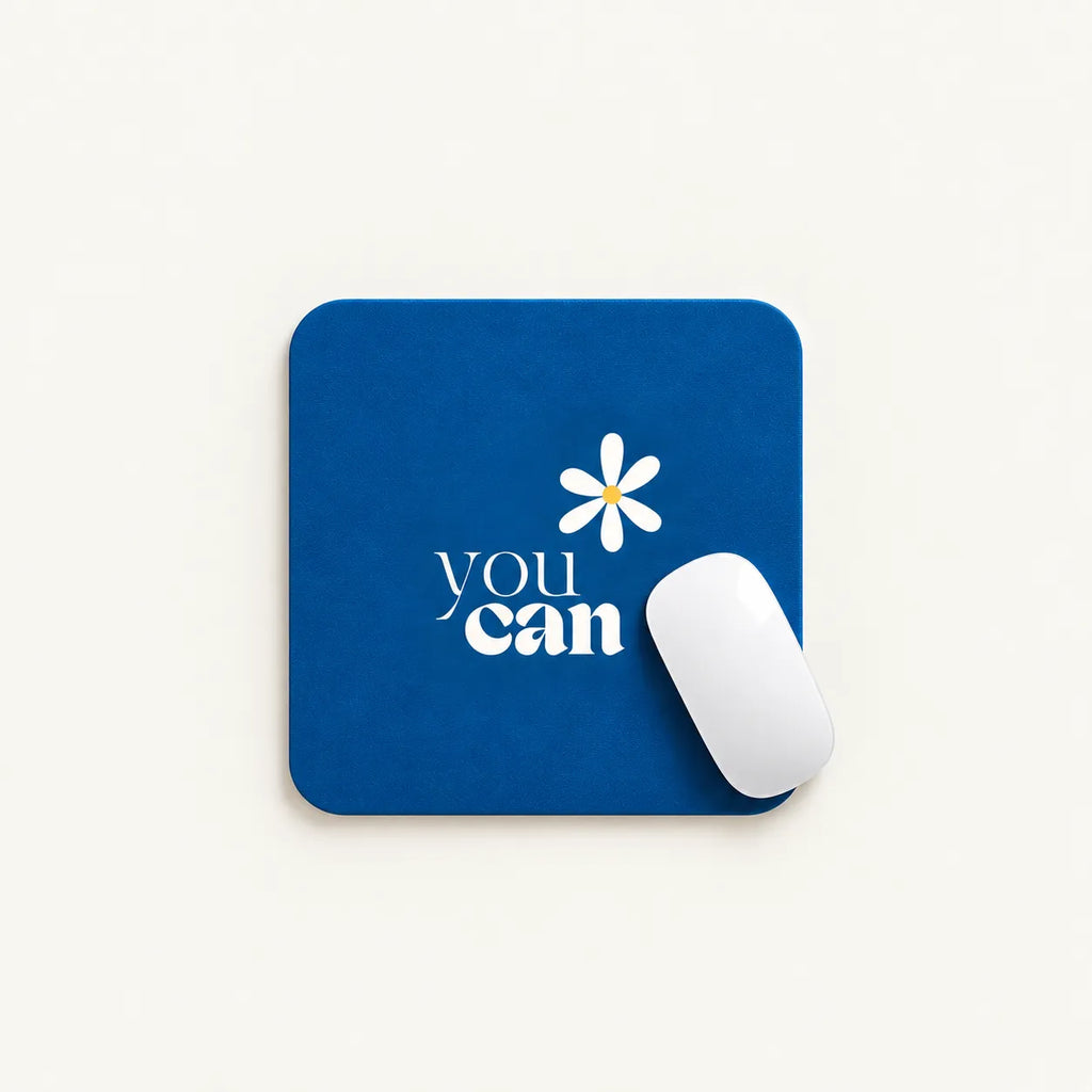 A square blue mouse pad with a white daisy graphic and the words 'you can' printed on it, shown on a light beige background with a white computer mouse resting on top.