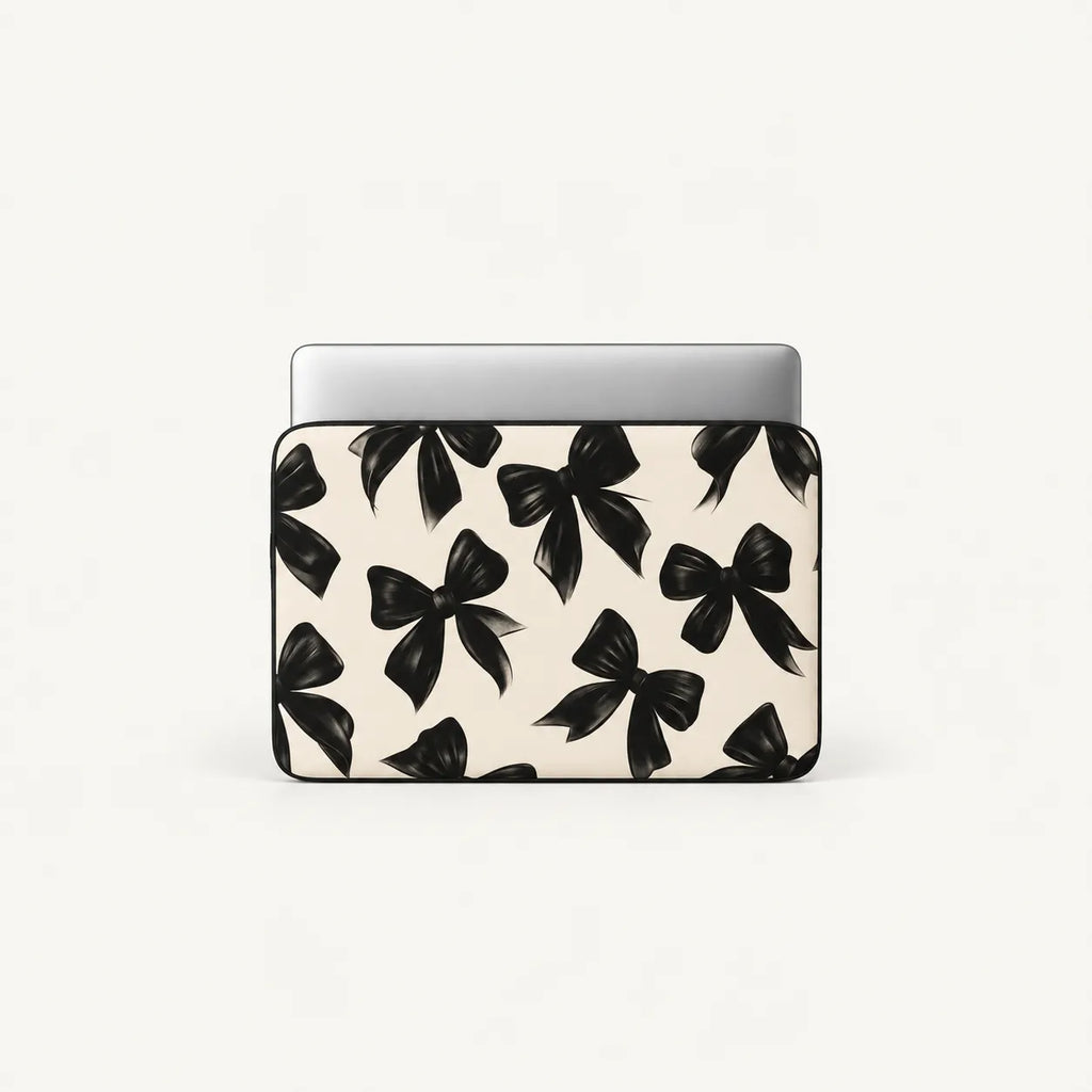 A laptop sleeve featuring a repeating black bow pattern on a warm off-white background, shown with a silver laptop inserted at the top, set against a plain light gray studio background.
