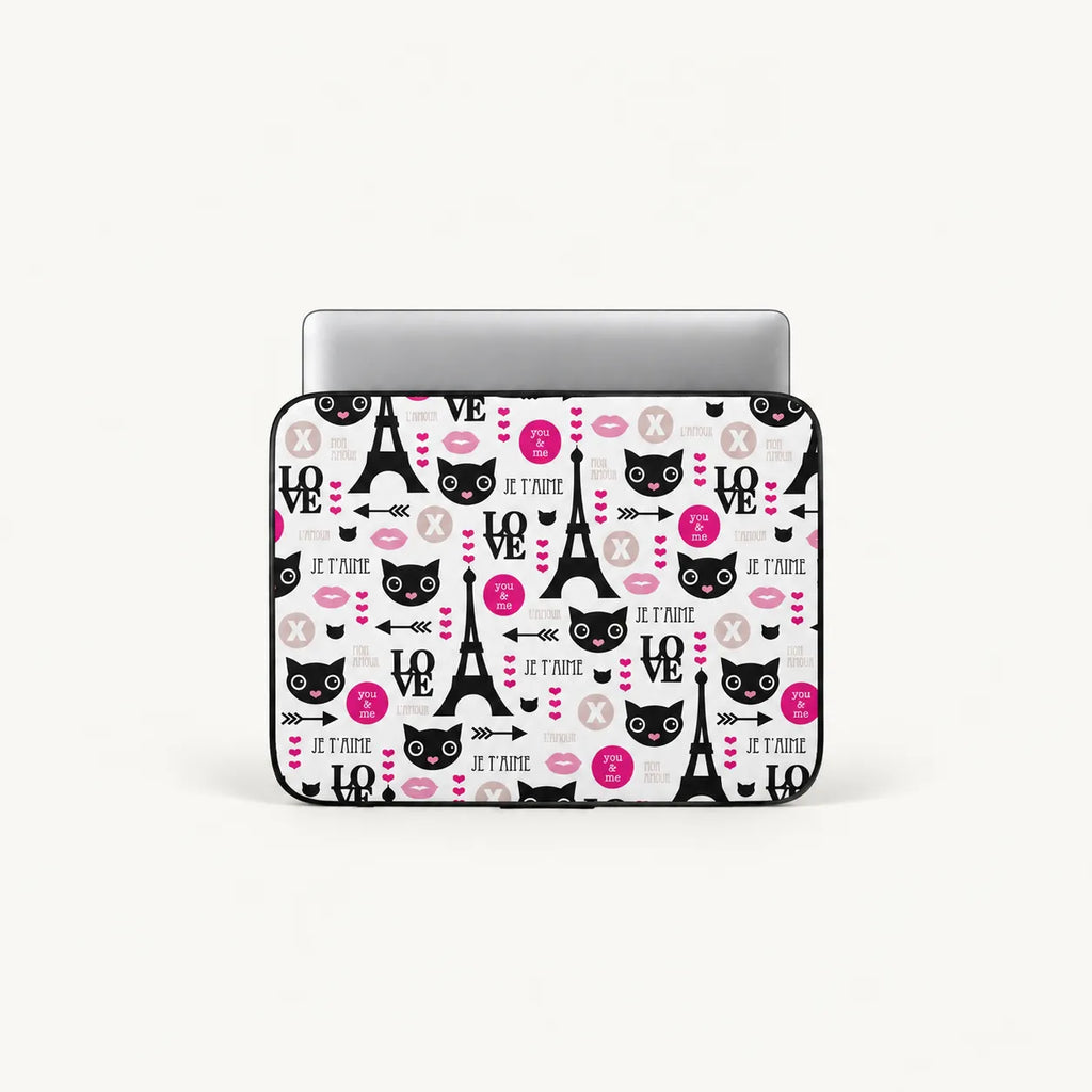 A laptop sleeve featuring a Paris-themed pattern with black cat faces, Eiffel Towers, pink hearts, and the phrase 'Je T'Aime' on a white background, shown with a silver laptop inserted.