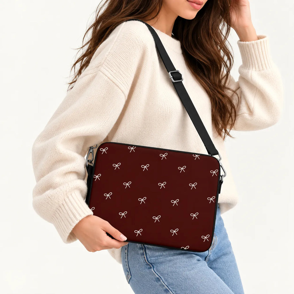 A woman wearing a cream knit sweater and light blue jeans carries a burgundy crossbody laptop bag with a repeating white bow pattern and a black strap.