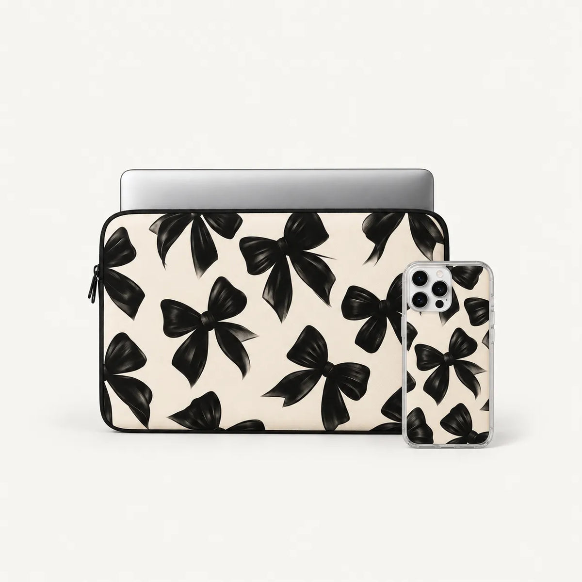 A laptop sleeve and matching phone case set featuring a black bow pattern on a warm cream background, shown on a clean off-white studio background.