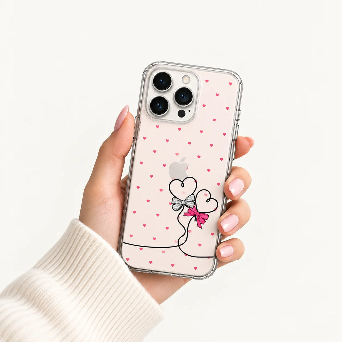 A hand holding a clear phone case with a pink background, small pink hearts, and a black-and-pink heart illustration near the bottom. The phone is partially visible inside the case, and the hand is wearing a cream ribbed sweater against a clean white background.