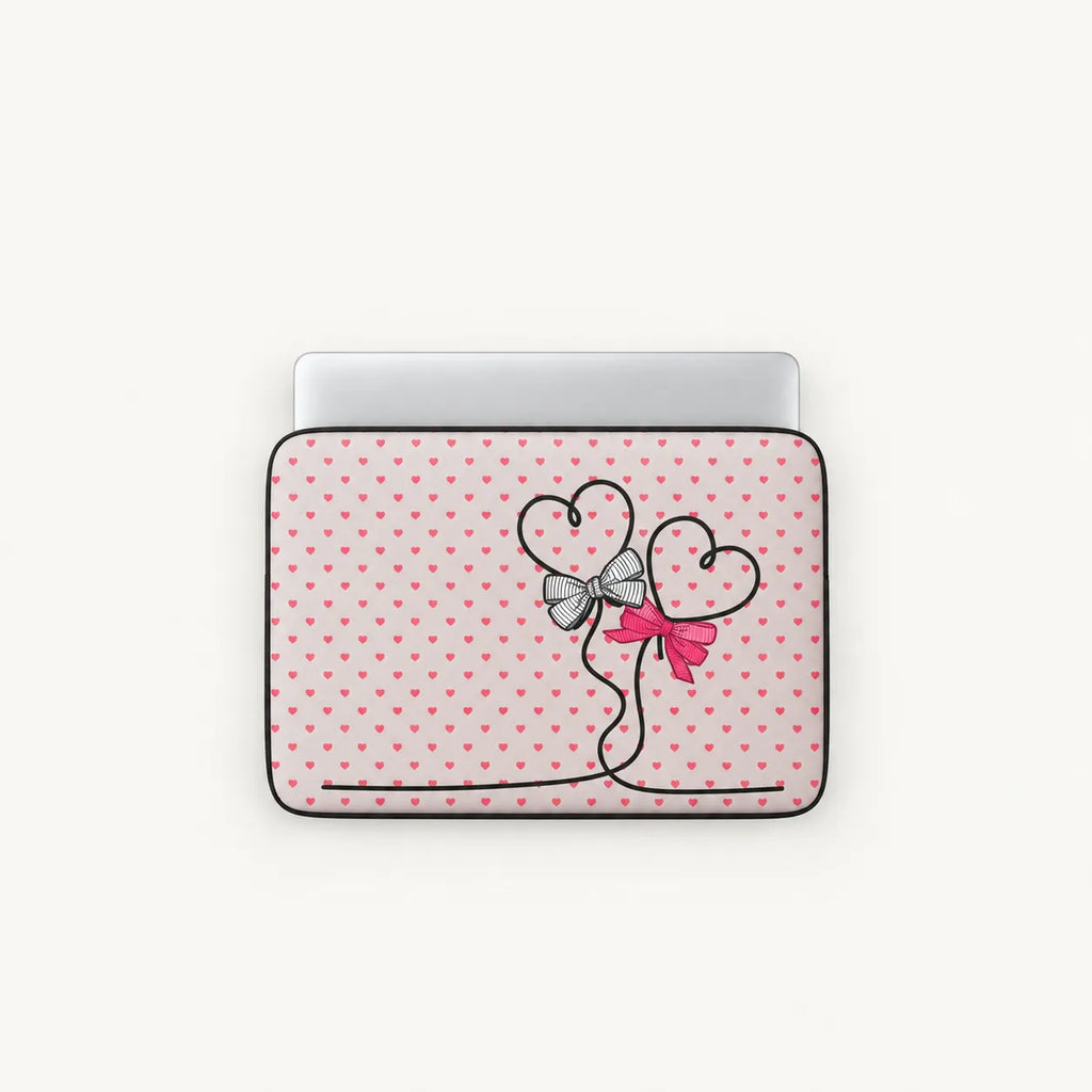 A laptop sleeve featuring a pink and white polka dot pattern with a black line-art heart design tied with a silver bow and a pink ribbon, shown on a light gray background.