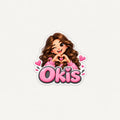 A die-cut sticker featuring a cartoon girl with wavy brown hair making a heart shape with her hands, above the word 'Okis' in pink bubble letters with heart details, on a light gray background.