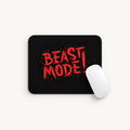 A black rectangular mouse pad with rounded corners featuring the words 'BEAST MODE!' in distressed red lettering, shown on a light gray background with a white computer mouse resting on the right side.