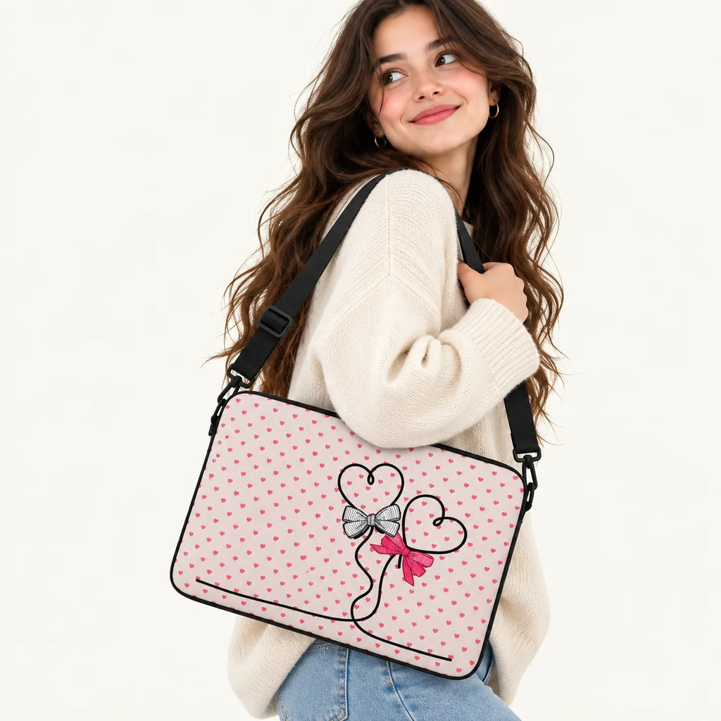 A woman wearing a cream knit sweater and blue jeans carries a pink laptop bag with a polka dot pattern and a heart-and-bow graphic on the front, set against a plain white background.