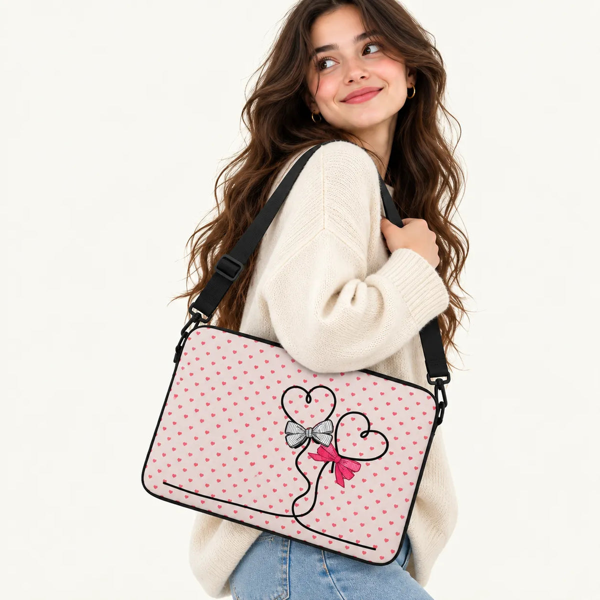 A woman wearing a cream knit sweater and blue jeans carries a pink laptop bag with a polka dot pattern and a heart-and-bow graphic on the front, set against a plain white background.