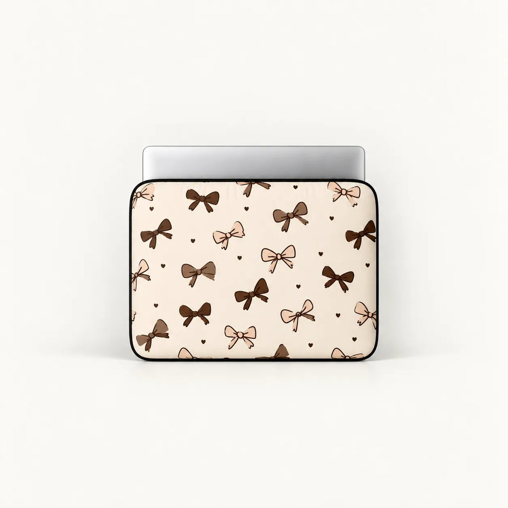 A laptop sleeve featuring a repeating pattern of brown and beige bows and small hearts on a cream background, shown with a silver laptop inside against a plain white background.