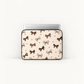 A laptop sleeve featuring a repeating pattern of brown and beige bows and small hearts on a cream background, shown with a silver laptop inside against a plain white background.