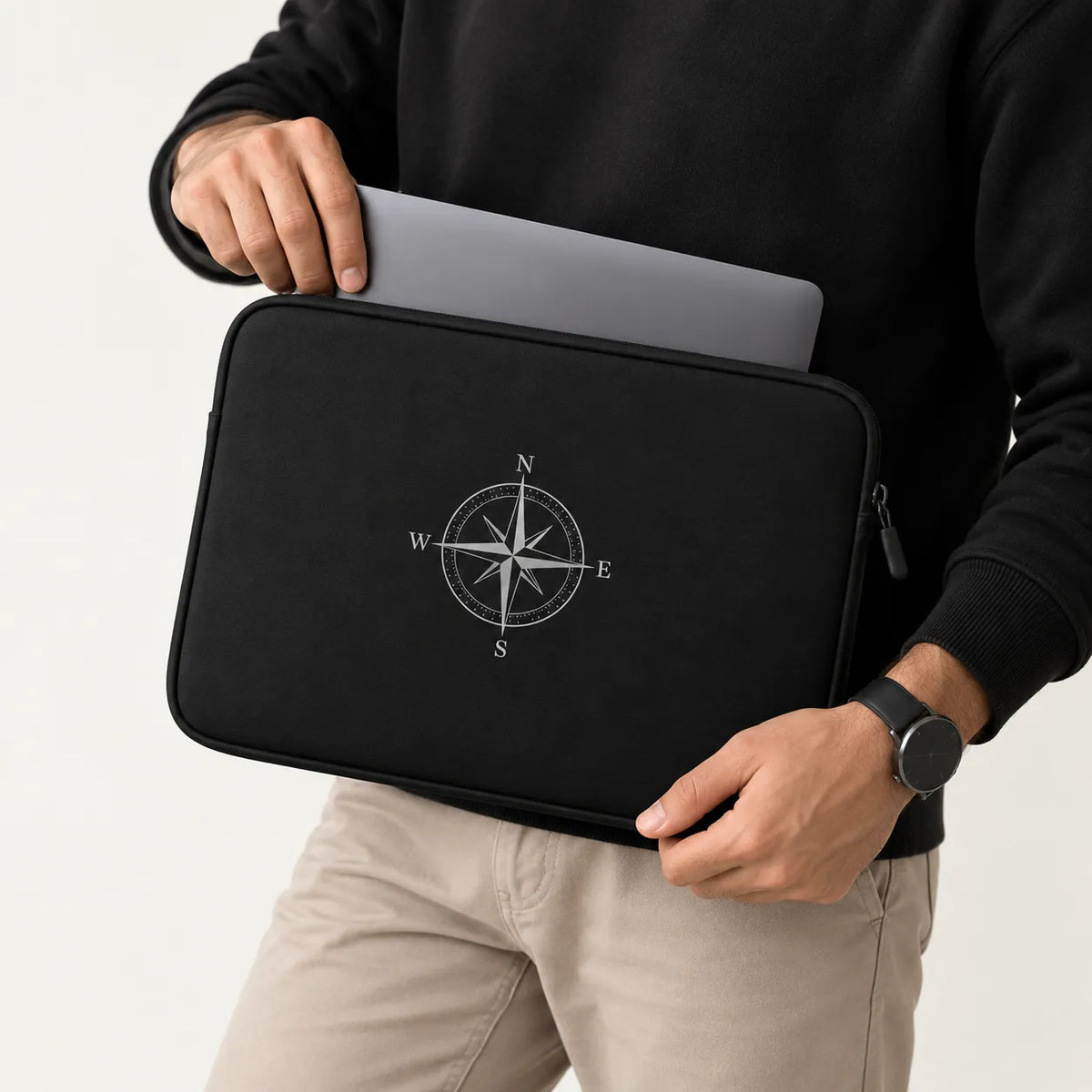 A person in a black sweatshirt and beige pants holds a black laptop sleeve with a white compass design. The sleeve is shown holding a gray laptop against a clean white background.