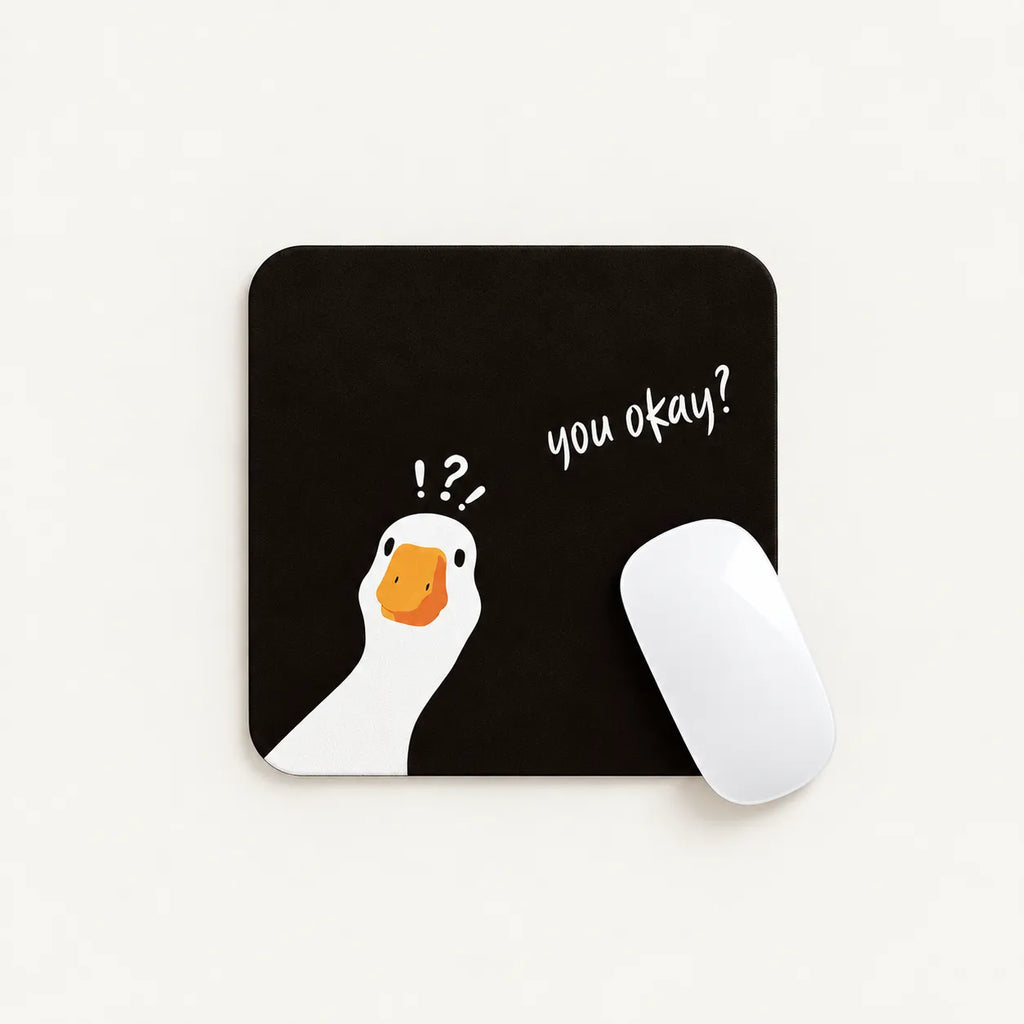 A square black mousepad with a white goose illustration, an orange beak, and the handwritten text 'you okay?' next to it. A white computer mouse rests on the right side of the pad, photographed on a clean off-white background.