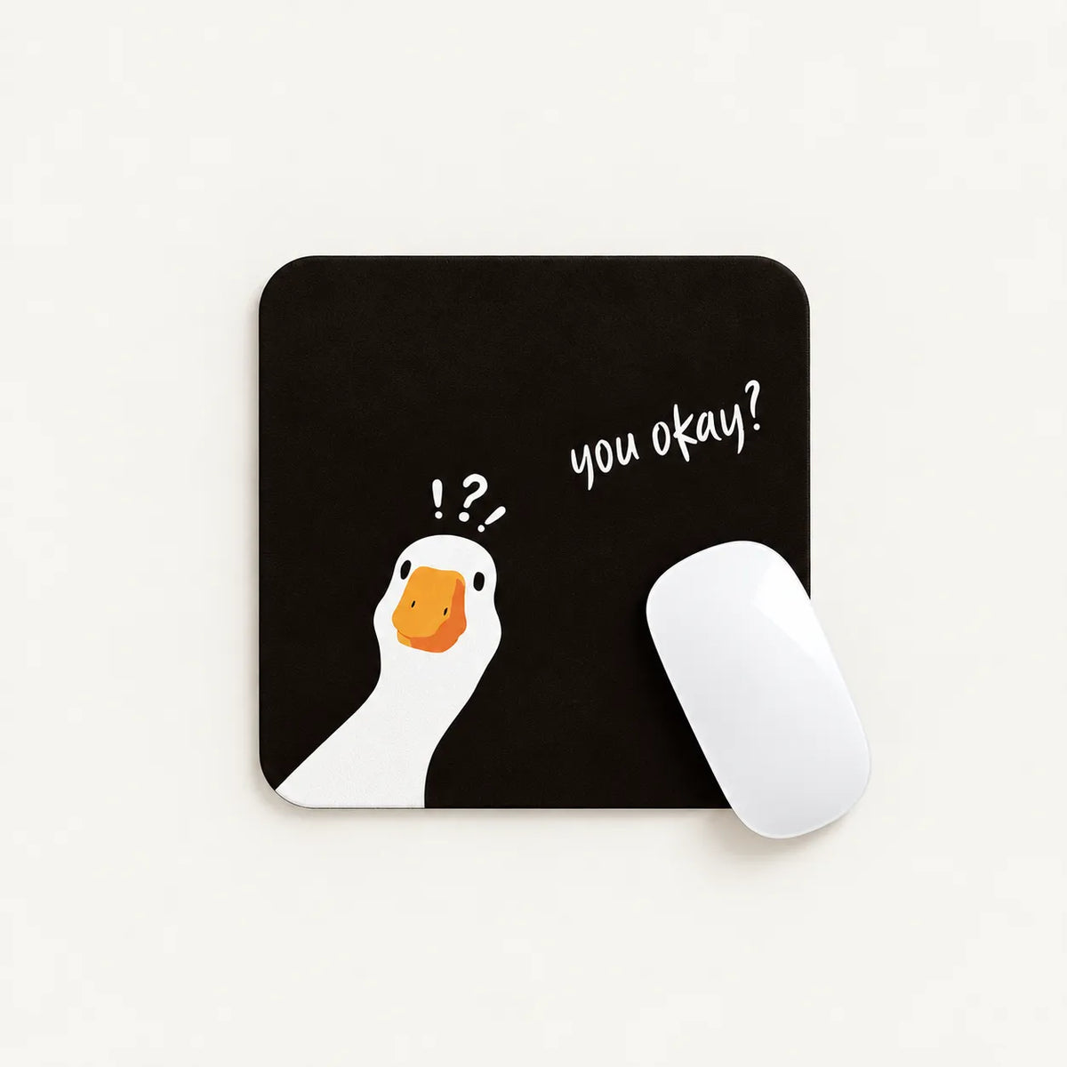 A square black mousepad with a white goose illustration, an orange beak, and the handwritten text 'you okay?' next to it. A white computer mouse rests on the right side of the pad, photographed on a clean off-white background.