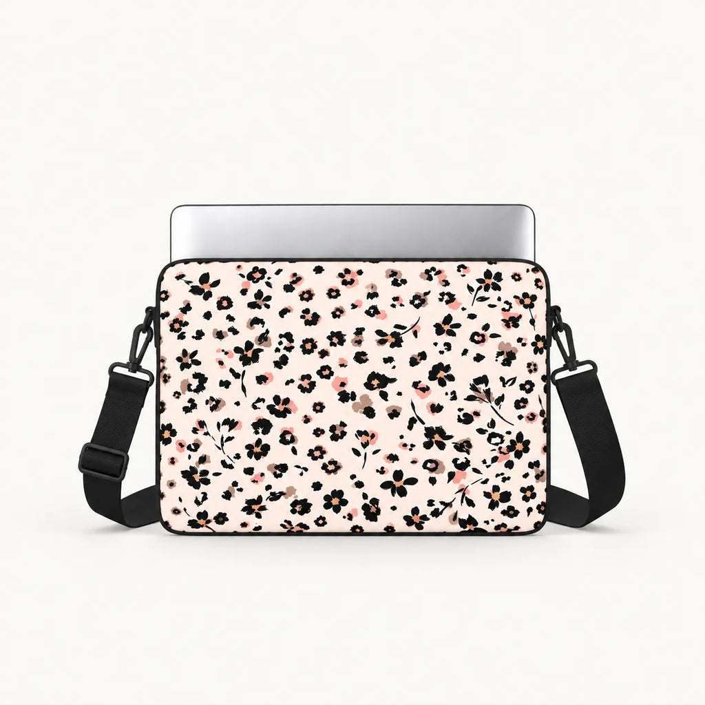A laptop sleeve with a black and pink floral pattern on a light pink background, shown with a silver laptop inserted and a black shoulder strap against a white studio background.