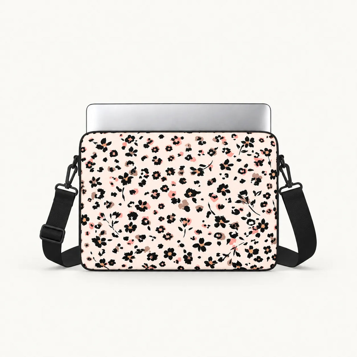 A laptop sleeve with a black and pink floral pattern on a light pink background, shown with a silver laptop inserted and a black shoulder strap against a white studio background.