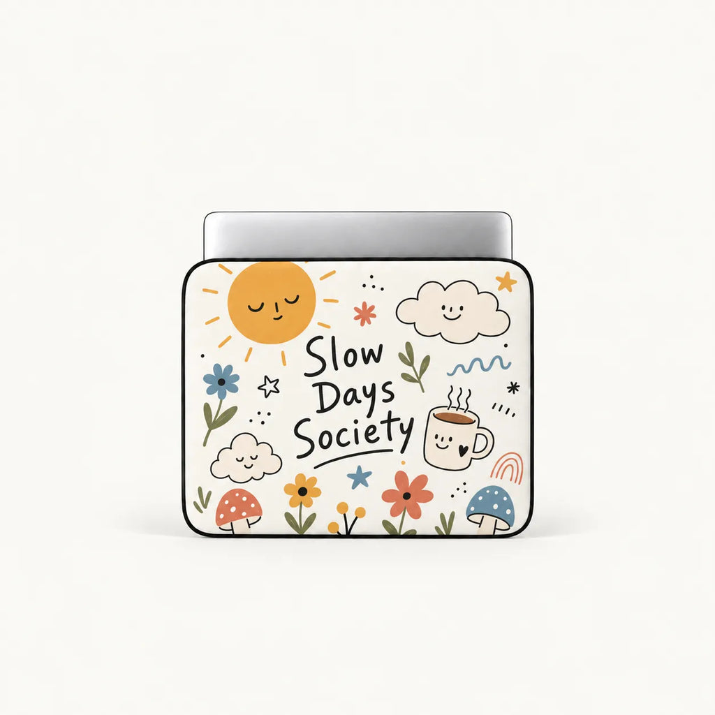 A laptop sleeve featuring a cute, hand-drawn style illustration with the text 'Slow Days Society'. The design includes a smiling sun, clouds, flowers, mushrooms, and a coffee cup on a white background with a black border.