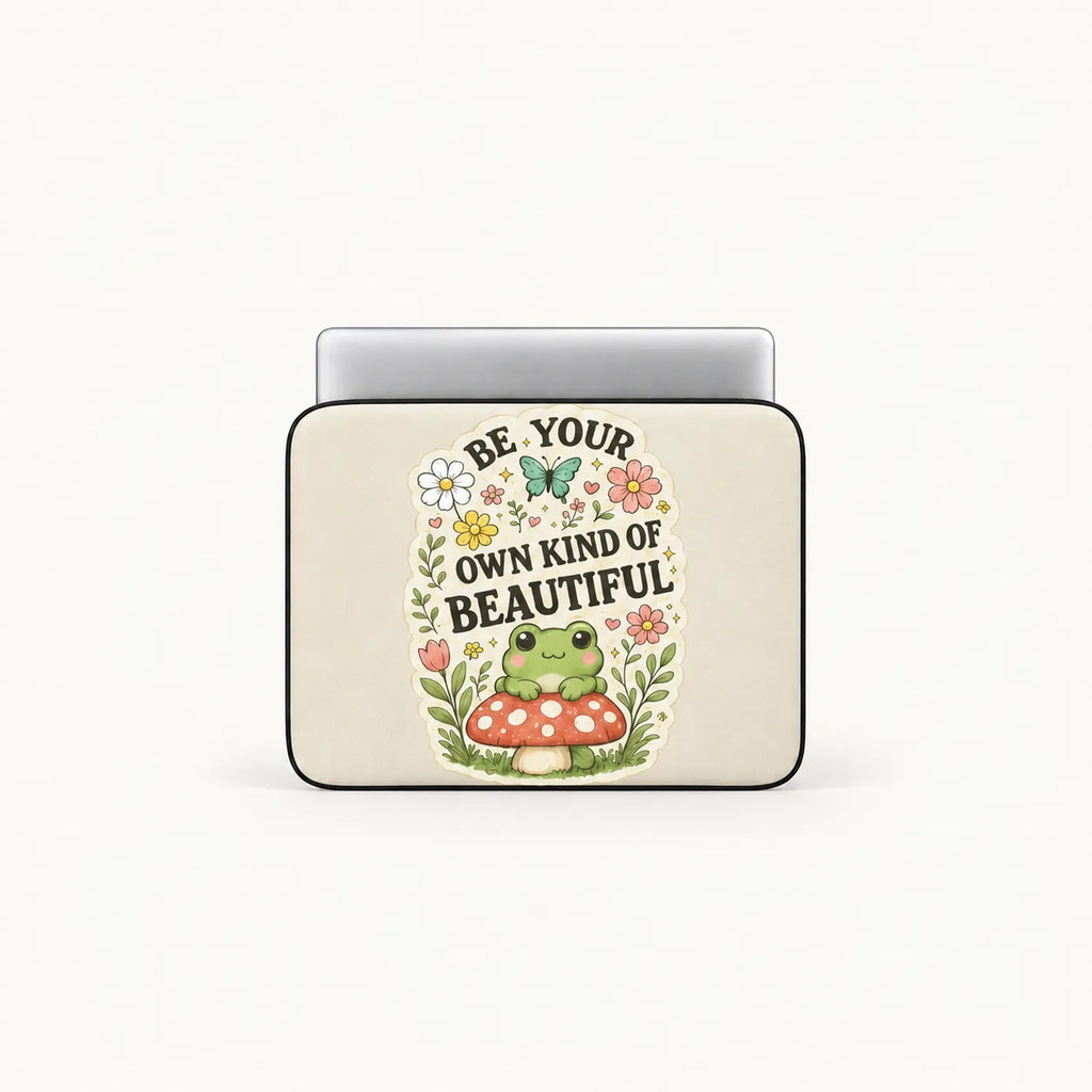 Cute green frog sitting on a red toadstool surrounded by flowers and Be Your Own Kind of Beautiful typography on cream padded laptop sleeve by SLEEVER