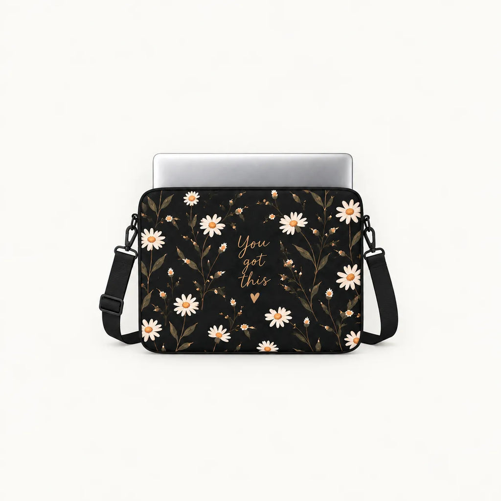 A black laptop sleeve with a white daisy and gold floral pattern, featuring the gold script text 'You got this' in the center. A silver laptop is partially inserted into the sleeve, which also has an adjustable black shoulder strap, set against a plain off-white background.