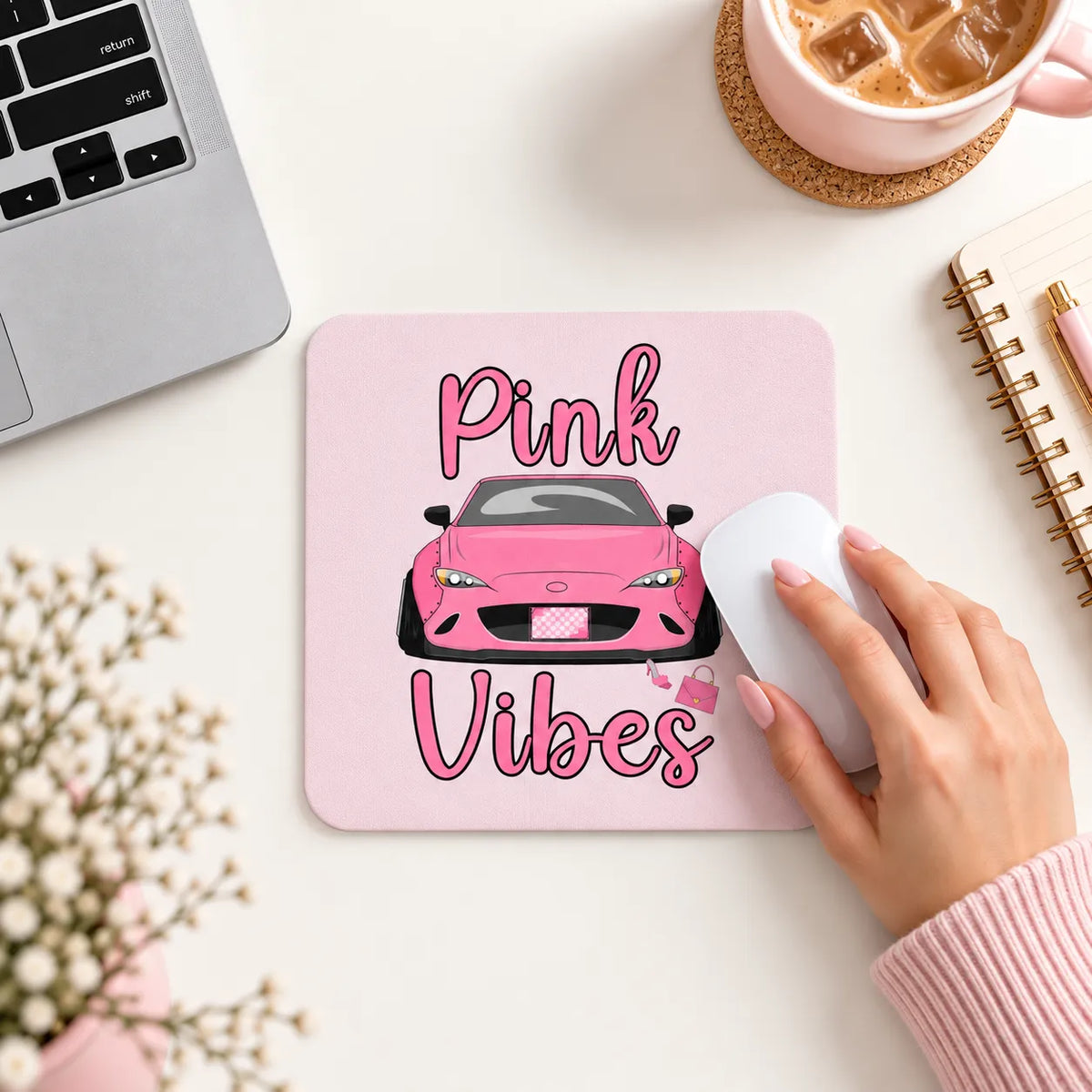 A pink rectangular mouse pad with a cartoon pink car and the words 'Pink Vibes' in pink script, shown on a white desk with a hand using a white mouse. A laptop corner, coffee cup, notebook, and flowers are visible around it.