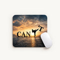 A square computer mouse pad with rounded corners, featuring a graphic design of a martial arts silhouette kicking a letter 'T' in a sunset sky, with the word 'CAN'T' on the pad. A white computer mouse is placed on the right side of the pad, set against a clean off-white background.