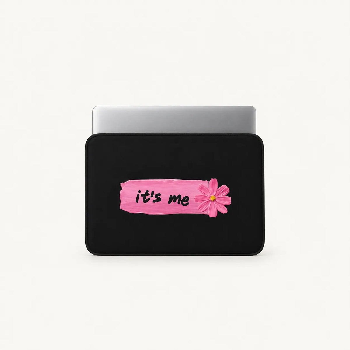 A black laptop sleeve with a pink paint-stroke graphic and a pink flower, featuring the text 'it's me' in black script, shown with a silver laptop on a light gray background.
