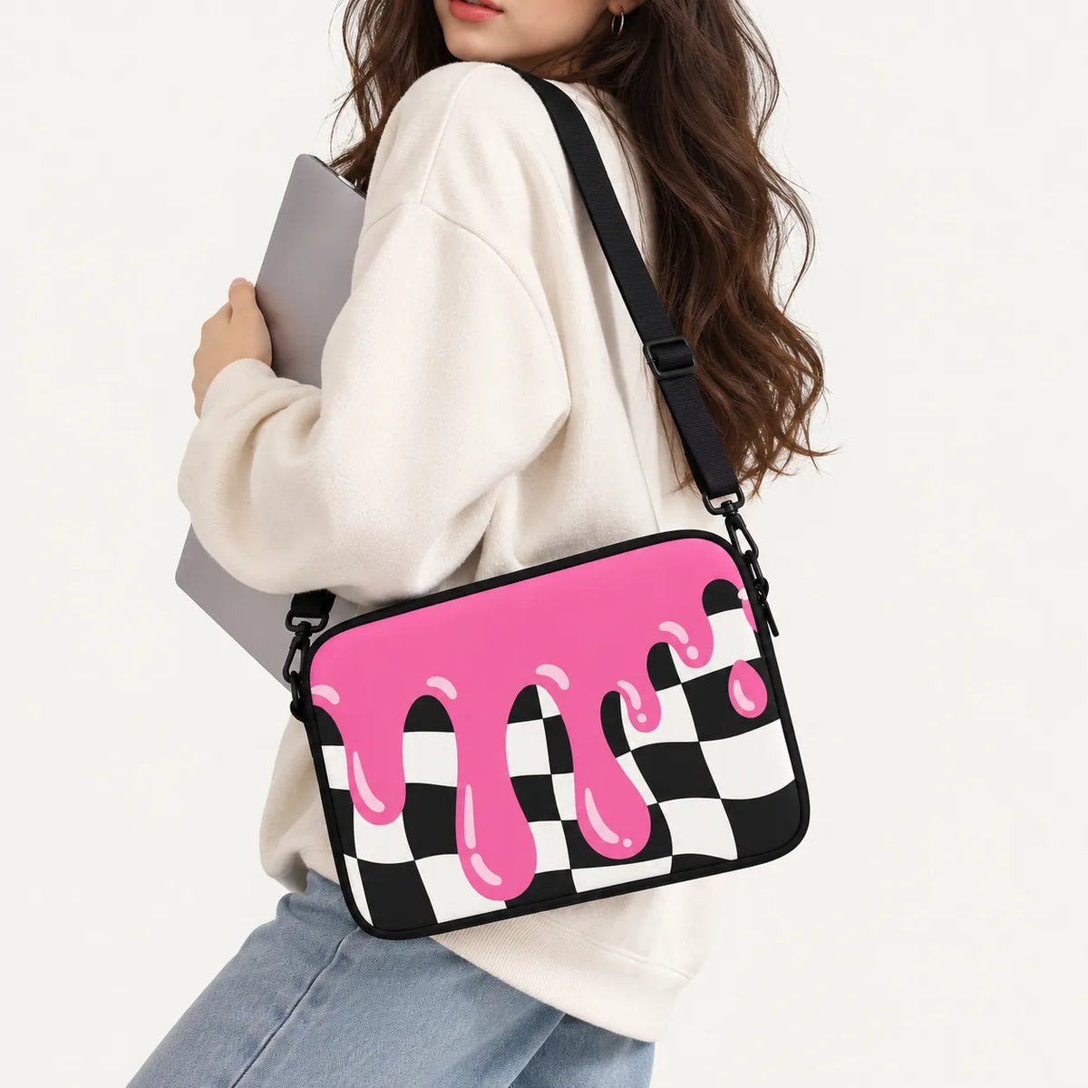 A woman wearing a cream sweatshirt and light blue jeans carries a black and white checkered crossbody bag with a bright pink dripping paint design on the front. She is holding a gray laptop against a clean white background.